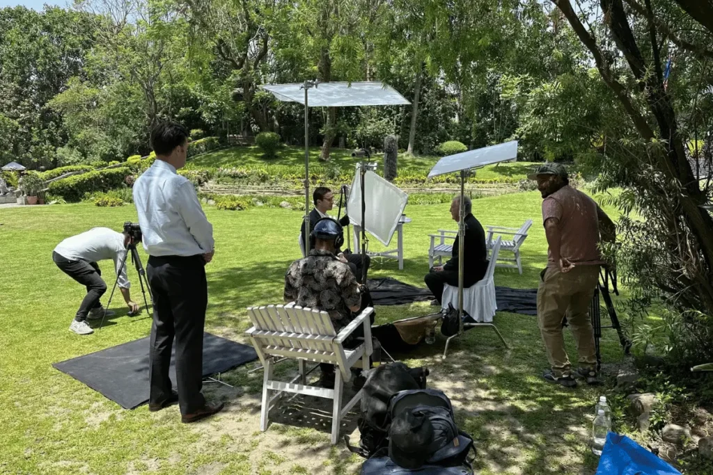 Local film crew setting up lighting reflectors and camera equipment for an outdoor interview shoot in a lush green garden in Nepal