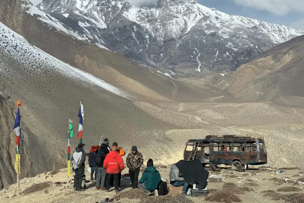 Film crew standing on a high-altitude mountain road in Nepal with snow-capped Himalayan peaks and colorful prayer flags in the background during a remote location shoot