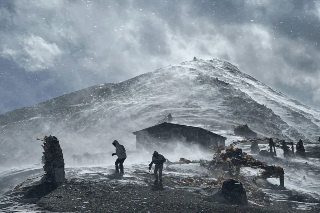 Two crew members pushing through a violent snowstorm while filming at Everest Base Camp, with prayer flags and a stone shelter barely visible through the whiteout conditions.