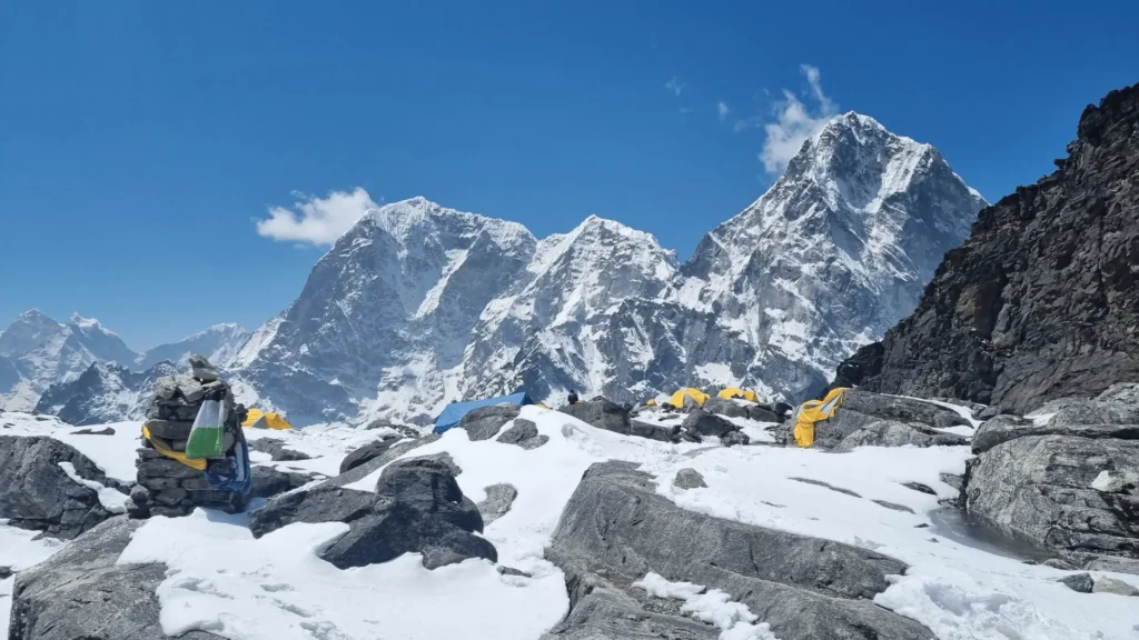 What no one tells you about filming at Everest base camp: Equipment, Altitude Sickness, and Sherpa Support Yellow expedition tents pitched on the Khumbu glacier moraine at Everest Base Camp, with a stone cairn wrapped in prayer flags in the foreground and the Khumbu Himalayan peaks rising under a clear blue sky.