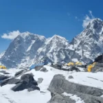 Yellow expedition tents pitched on the Khumbu glacier moraine at Everest Base Camp, with a stone cairn wrapped in prayer flags in the foreground and the Khumbu Himalayan peaks rising under a clear blue sky.