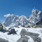 Yellow expedition tents pitched on the Khumbu glacier moraine at Everest Base Camp, with a stone cairn wrapped in prayer flags in the foreground and the Khumbu Himalayan peaks rising under a clear blue sky.