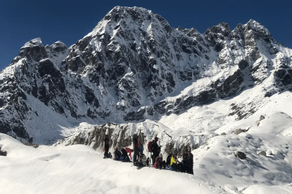 Film crew resting on a glacial snowfield during high altitude filmmaking in Nepal, with a dramatic snow-covered Himalayan peak rising against a deep blue sky.