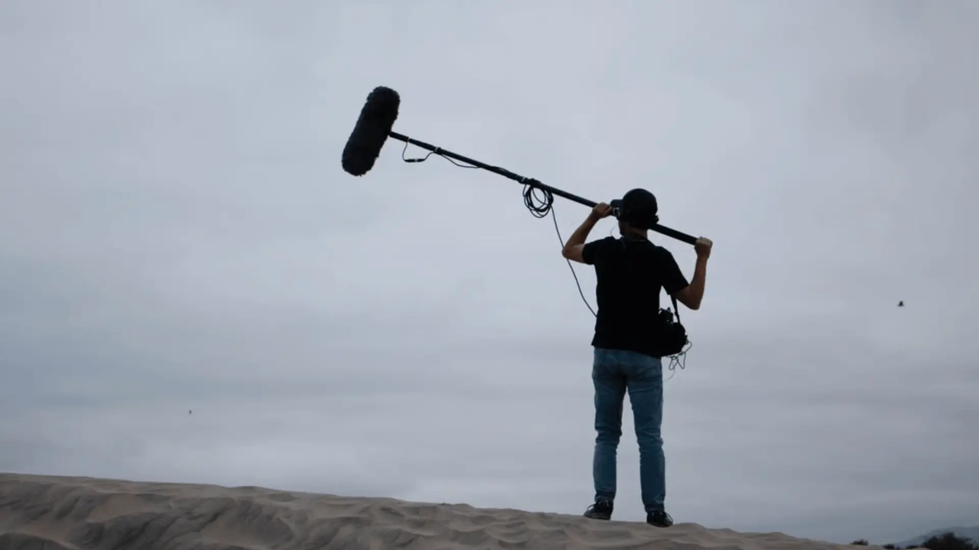 Boom operator recording location sound with a windshielded mic on a ridge.